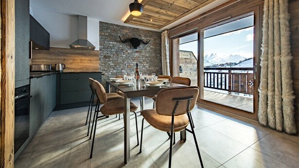 Une cuisine moderne avec un mur de pierres apparentes derrière le plan de travail, un coin repas avec une table dressée pour quatre personnes et une grande fenêtre offrant une vue sur les montagnes.