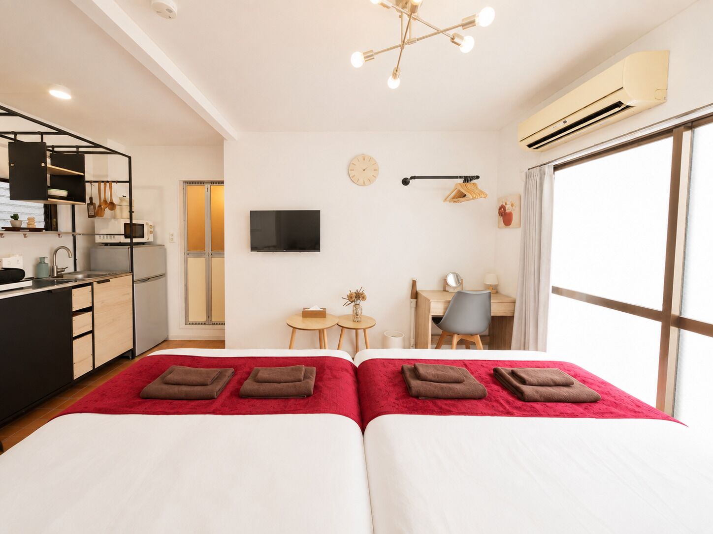 A hotel room with two beds, a kitchenette, a desk, a chair, a TV, and a large window.