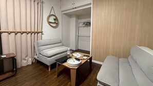A compact room with a sofa, a small table with a clock, a shelf with a plant, and a wardrobe. - NEST BASE IKEBUKURO (Tokyo)