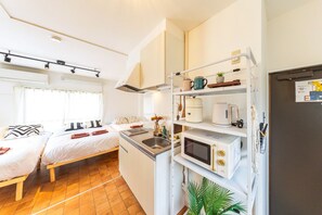 A compact kitchenette with a microwave, kettle, and small fridge. There is a bed with pillows and a window with curtains. - NEST BASE IKEBUKURO (Tokyo)