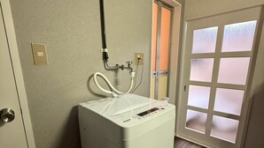 A compact laundry room with a front-loading washing machine, a hose, and a door with frosted glass panels. - NEST BASE IKEBUKURO (Tokyo)