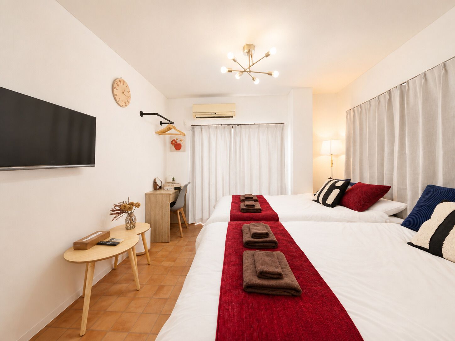 A hotel room with two beds, a desk, a chair, a TV, a clock, and a ceiling light fixture.