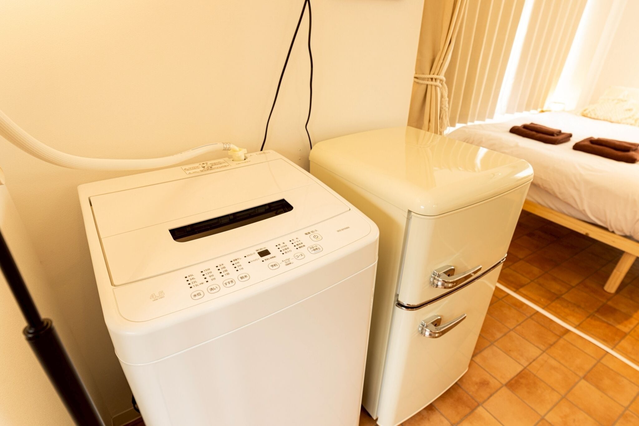 A compact, white laundry machine with a control panel and a small drawer unit, situated in a room with a bed and a curtain.