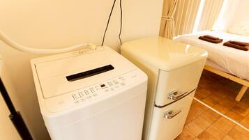 A compact, white laundry machine with a control panel and a small drawer unit, situated in a room with a bed and a curtain.