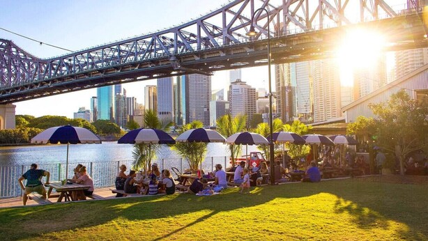 A riverside park with picnic tables and umbrellas, a bridge in the background, and a city skyline.