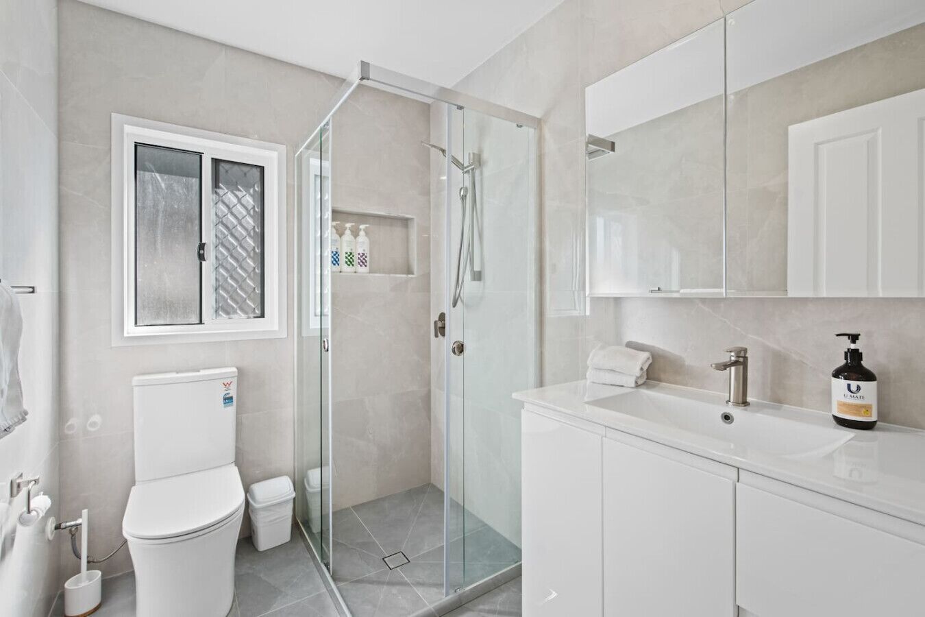 Une salle de bain moderne avec une douche à parois de verre, des toilettes, un lavabo avec un miroir et un flacon de savon U-Matt.