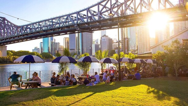 Un parque junto al río con mesas de picnic y sombrillas, un puente al fondo y el perfil urbano de la ciudad.
