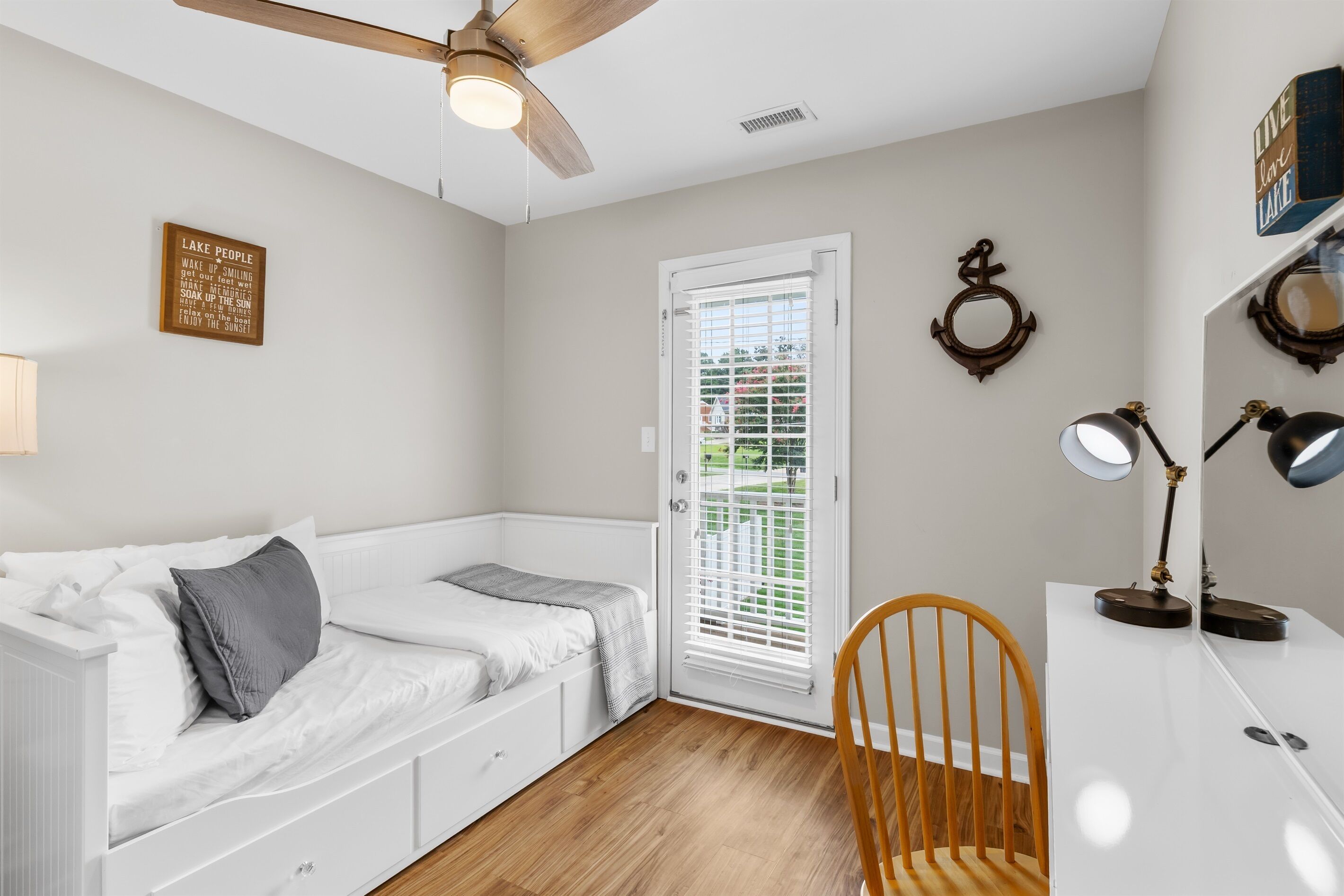 Une chambre à coucher comprenant un lit, un bureau, une chaise, un ventilateur de plafond, une fenêtre avec des stores et un panneau affiché sur le mur.