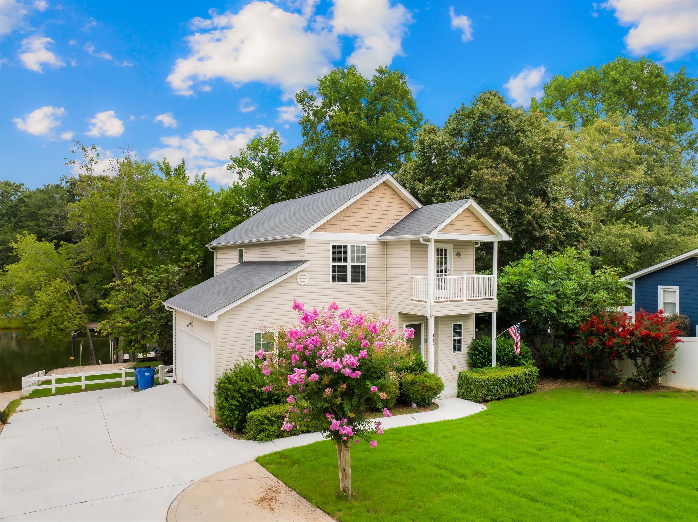 A two-story house with a front porch and a small deck, surrounded by lush greenery and a clear blue sky.