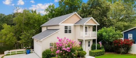 A two-story house with a front porch and a small deck, surrounded by lush greenery and a clear blue sky.
