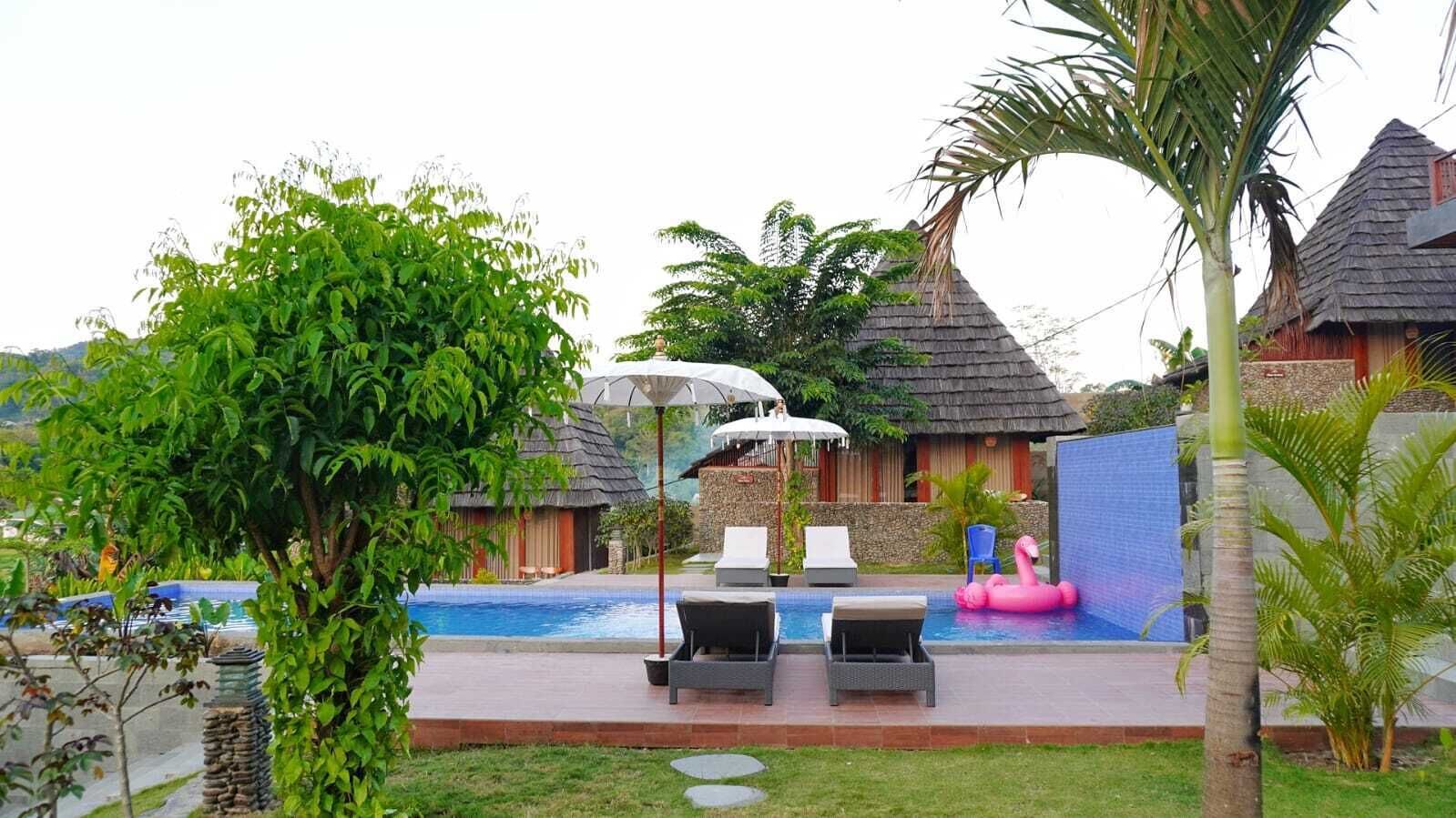 Un área de piscina con sillones de descanso y sombrillas, rodeada de plantas tropicales y edificios con techo de paja.
