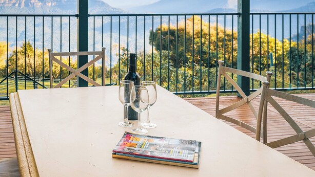 A table with a magazine, a bottle, and three glasses on a balcony with a mountain view. - Blue Mountains Rhapsody - Point Blank Views Clifftop (Leura)