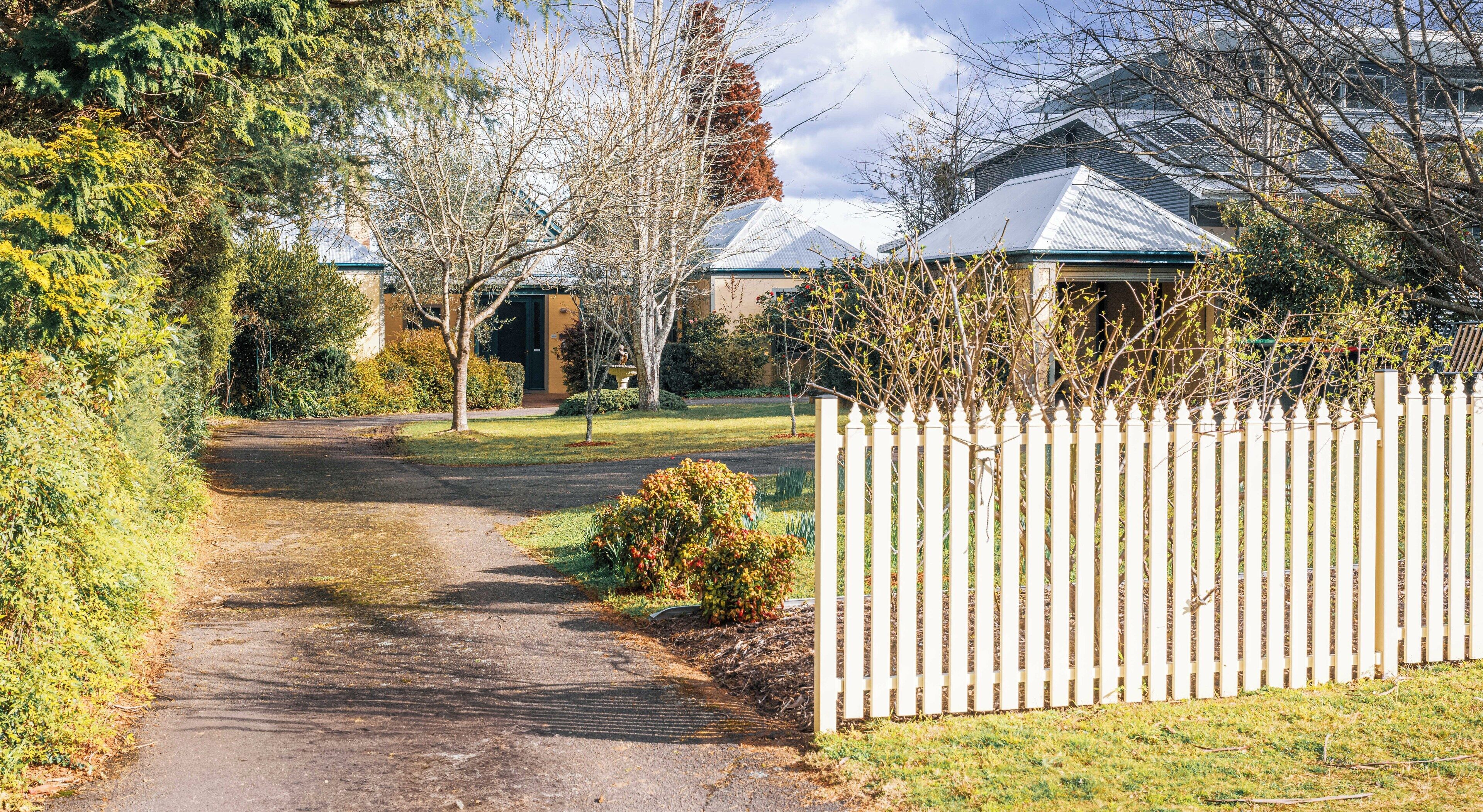 A pathway leading to a gated area with a white picket fence, surrounded by greenery and trees.