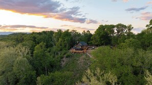 Et stort, tosset hus omgivet af tæt skov, med en fremtrædende terrasse og et stort vindue med udsigt over landskabet.
