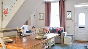 Ein gemütlicher Essbereich mit einem Holztisch, Stühlen und einer Bank. Im Hintergrund befindet sich eine Couch mit Kissen und eine Tür mit Fußmatte.