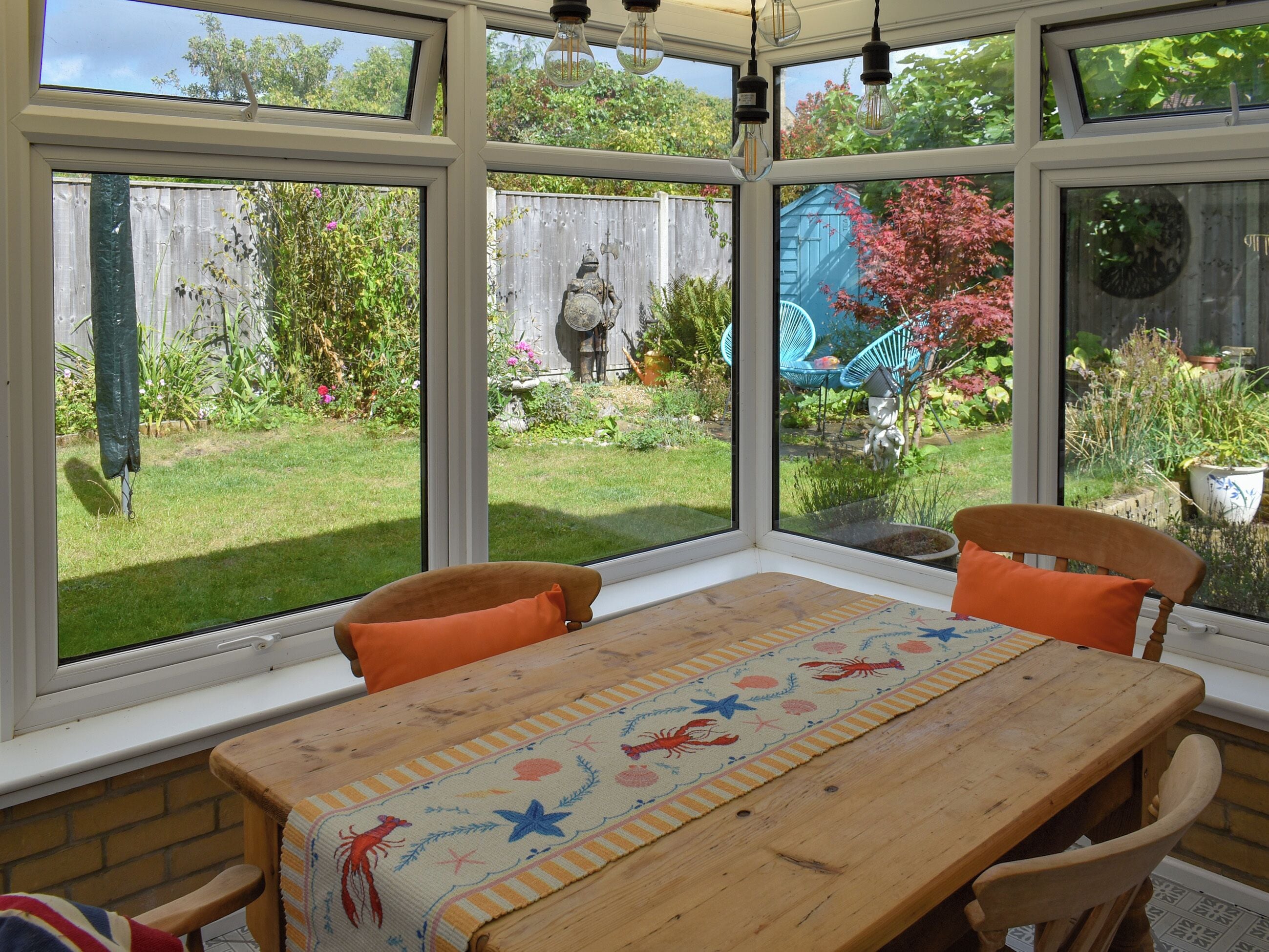 A conservatory with a wooden table, two chairs, and a view of a garden with a wooden fence and various plants.