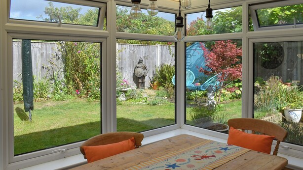 A conservatory with a wooden table, two chairs, and a view of a garden with a wooden fence and various plants. - 2 bedroom accommodation in Pakefield, near Lowestoft (Pakefield, near Lowestoft)