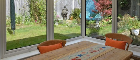 A conservatory with a wooden table, two chairs, and a view of a garden with a wooden fence and various plants.
