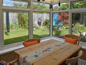 Un invernadero con una mesa de madera, dos sillas y vistas a un jardín con cerca de madera y diversas plantas.