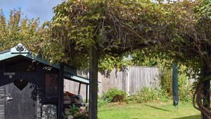 Un abri de jardin en bois orné d’une découpe en forme de cœur, doté d’un petit porche et d’un espace jardin avec de l’herbe et des plantes.