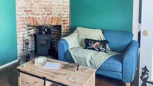 Un salon chaleureux avec une cheminée en briques, un canapé bleu agrémenté d’un plaid vert et une table basse en bois sur laquelle sont posés des livres et des télécommandes.