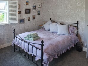 Una cama bien hecha con estructura metálica, una ventana con contraventanas y un estampado floral en el papel tapiz.