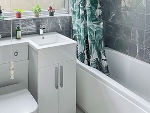 Un baño moderno con un lavabo blanco, un inodoro y una bañera. Hay plantas en macetas en el alféizar de la ventana.