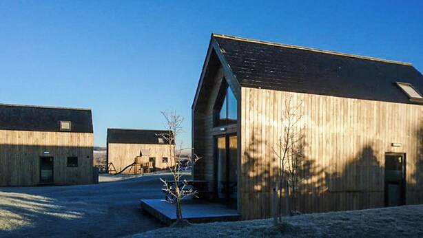 A modern building with a large glass entrance and a sloped roof, surrounded by other similar structures. - Modern comfort meets rugged wilderness at Tarset Tor’s unique glamping retreat. (Hexham, near Bellingham)