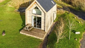 A small wooden cabin with a sloped roof, a glass door entrance, and a patio with a table and chairs. - Modern comfort meets rugged wilderness at Tarset Tor’s unique glamping retreat. (Hexham, near Bellingham)