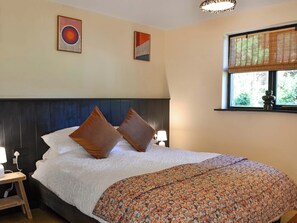 Een hotelkamer met een bed, twee kussens, een nachtkastje met een lamp, een raam met jaloezieën en twee kunstwerken aan de muur.