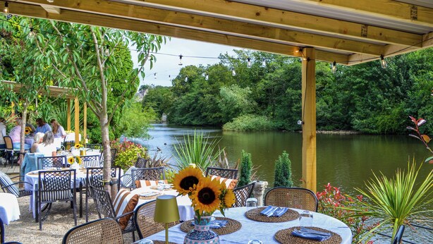 An outdoor dining area with tables set for two, surrounded by lush greenery and a river view.