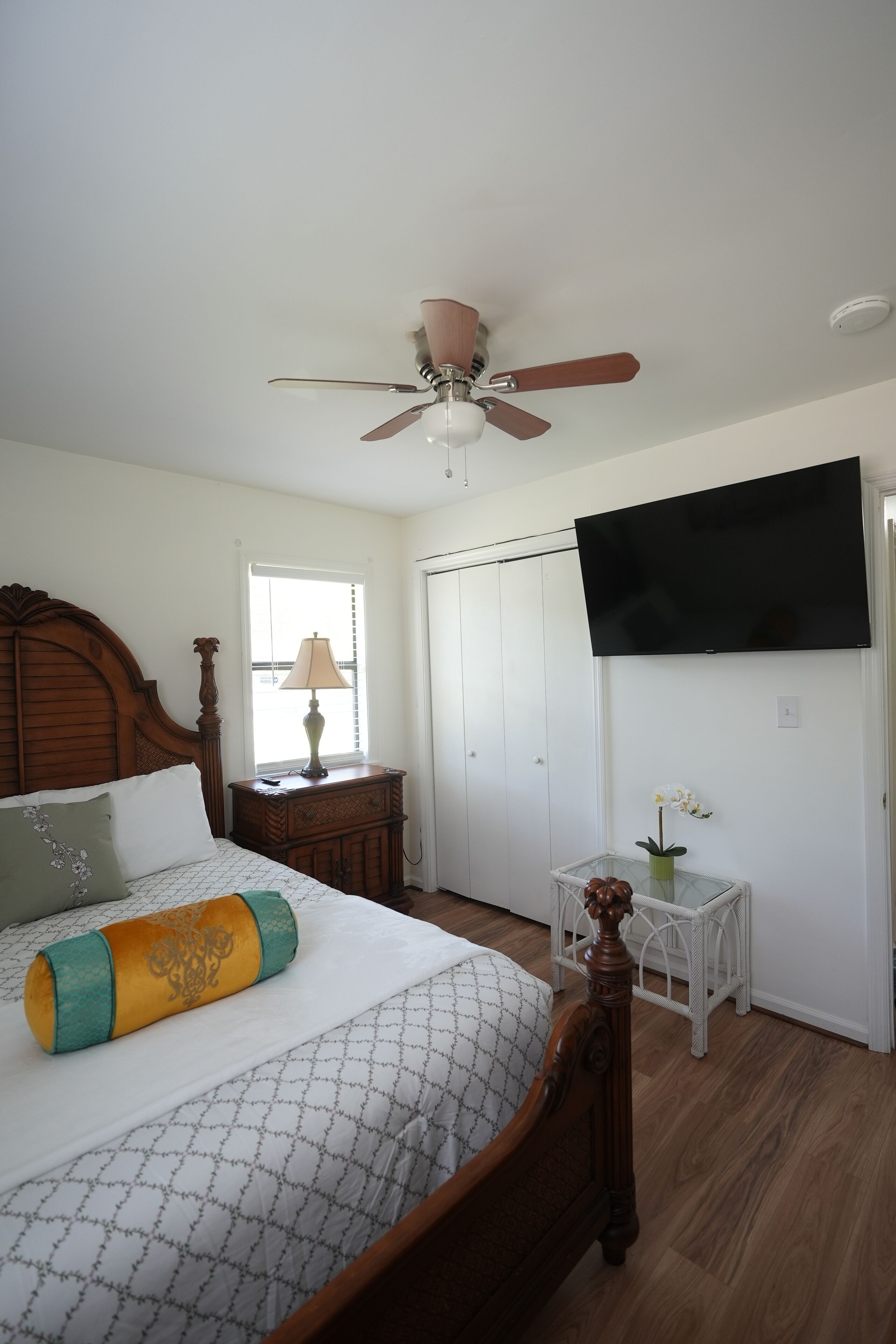Une chambre à coucher avec un lit, un ventilateur de plafond, une télévision, une fenêtre et une petite table avec une lampe.