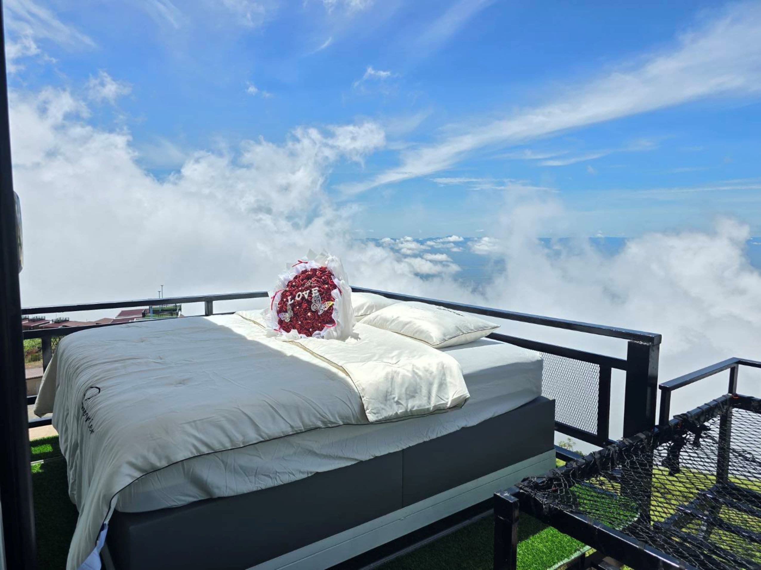A bed with a white bedspread and pillows, a bouquet of red roses, and a view of clouds and a distant landscape.