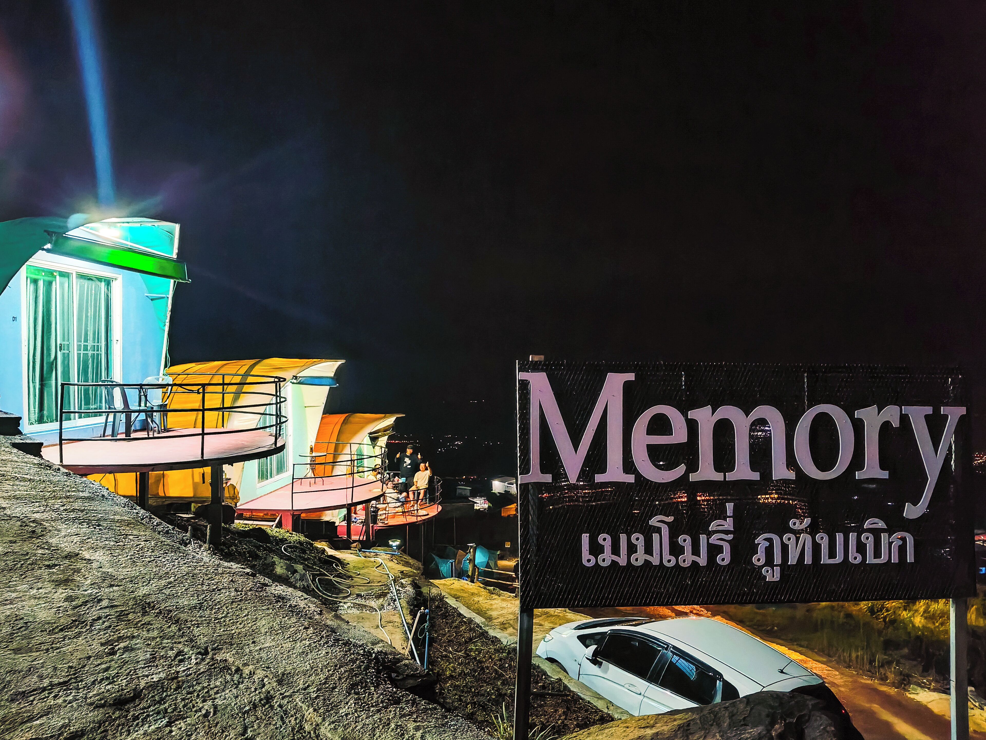 Ein Schild mit der Aufschrift "Memory" und thailändischem Text, ein Gebäude mit farbenfroher Fassade und ein Auto auf einer Straße.