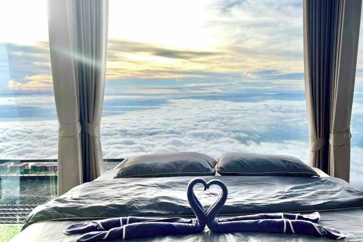 Una cama con una toalla dispuesta en forma de corazón, vista de nubes y un paisaje lejano a través de amplias ventanas.