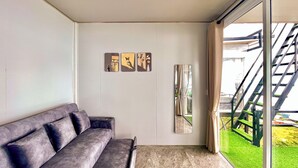 A modern living room with a grey sofa, a large window with a view of a green garden and a staircase, and three framed artworks on the wall.
