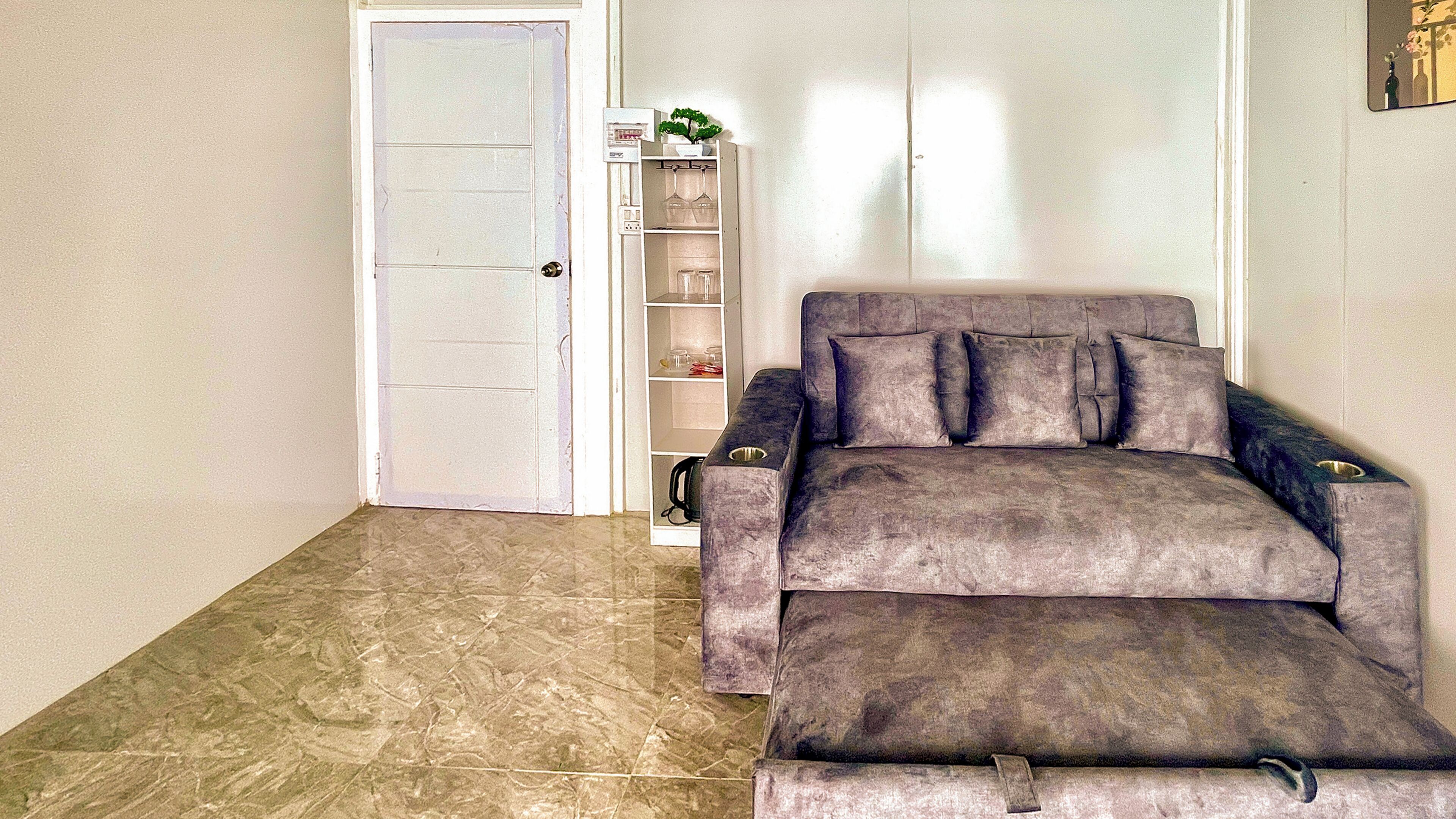 Una habitación con un sofá gris, una puerta blanca y una estantería blanca con cristalería.