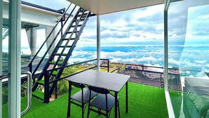 A balcony with a table and chairs, a staircase, and a view of clouds and buildings below.