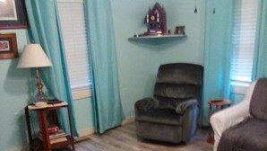 Un salon avec un plancher en bois, un fauteuil gris, un canapé, une petite table avec une lampe et une étagère fixée au mur.