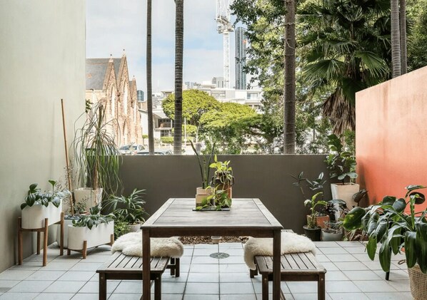 A balcony with a wooden table and benches, surrounded by potted plants and a city view.