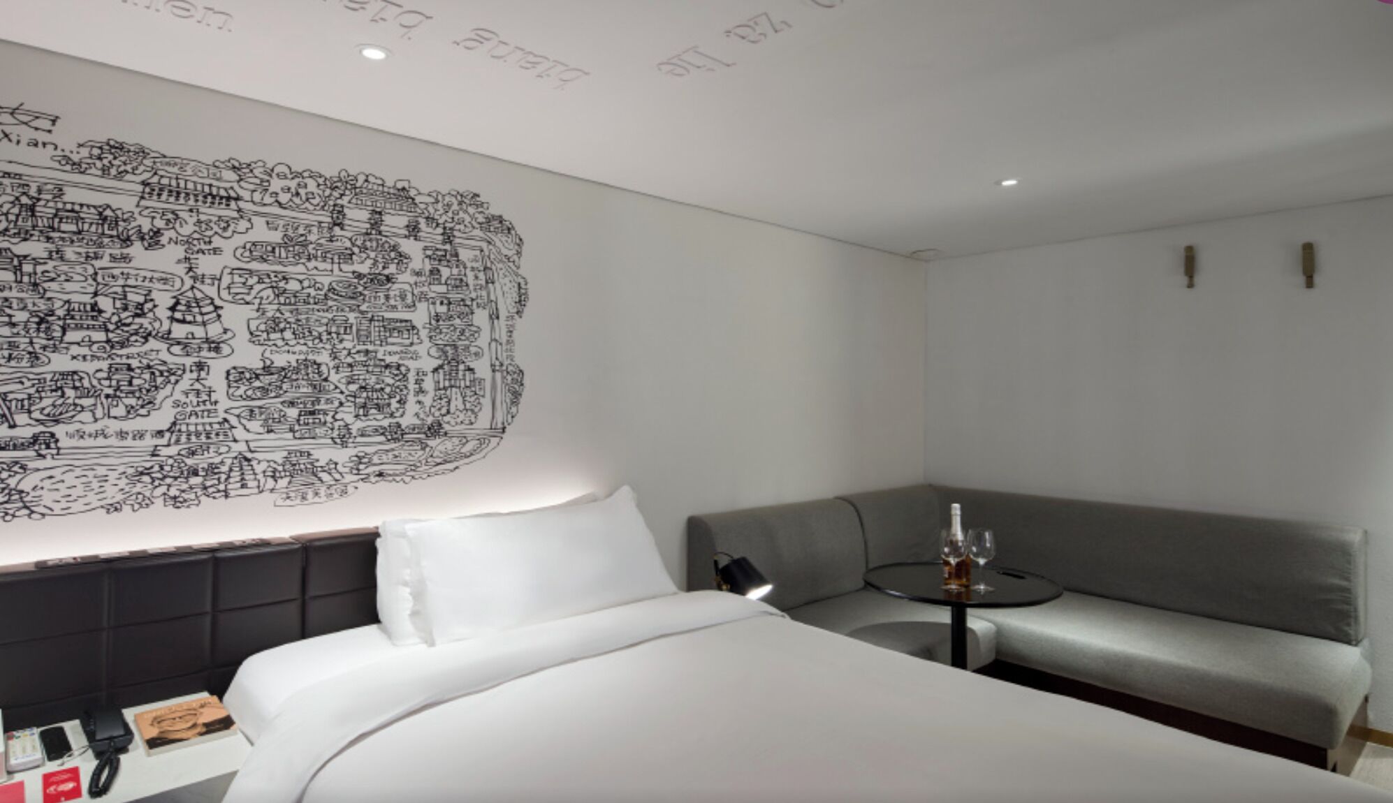 A hotel room with a bed, a sofa, a small table with a bottle, and a wall with a black and white cityscape mural.