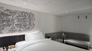 Une chambre d’hôtel avec un lit, un canapé, une petite table sur laquelle se trouve une bouteille, et un mur orné d’une fresque représentant un paysage urbain en noir et blanc.