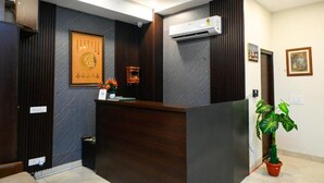 A reception area with a dark wood reception desk, a framed symbol on the wall, an air conditioning unit, and a potted plant. - HOTEL INFRAVIEW (Dera Bassi)
