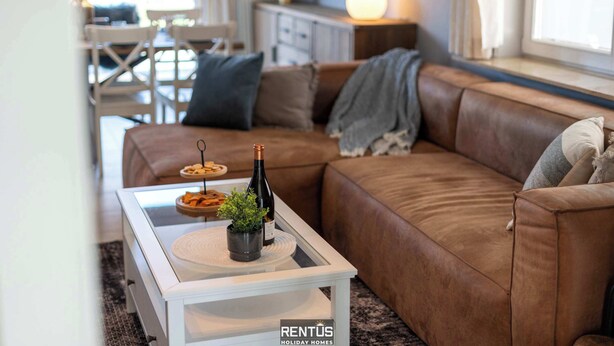 Una sala de estar acogedora con un sofá de cuero marrón, una mesa de centro blanca con una botella y una planta pequeña, y un comedor al fondo.