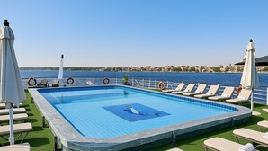 Piscina rectangular al aire libre con azulejos azules, rodeada de sillones de descanso y sombrillas, con vistas a una masa de agua y una orilla distante.