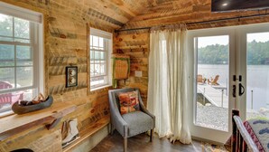 Habitación tipo cabaña de madera con techo de madera, un ventanal grande con vistas a un lago, una silla con cojín y una pequeña mesa con un cuenco de piñas.