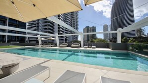 Una piscina moderna al aire libre con sillas de descanso y sombrillas, rodeada de edificios altos.