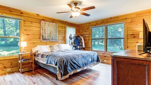 Un dormitorio tipo cabaña de madera con cama, ventilador de techo y ventanas amplias.