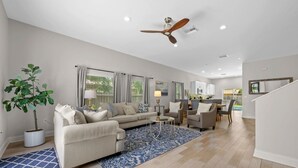 A spacious living room with a sofa, coffee table, and a large blue rug. There is a ceiling fan, a potted plant, and a view into the kitchen area.