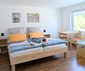 A neatly arranged bedroom with a wooden bed, two pillows, a bedside table, a chair, a small table, and a window.